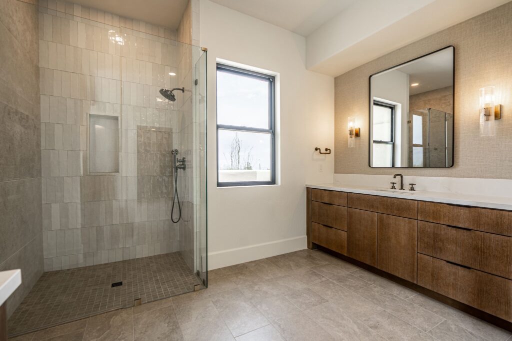 Modern bathroom with a glass-enclosed shower, a large window, dual-sink wooden vanity, rectangular mirror, and wall-mounted lights.