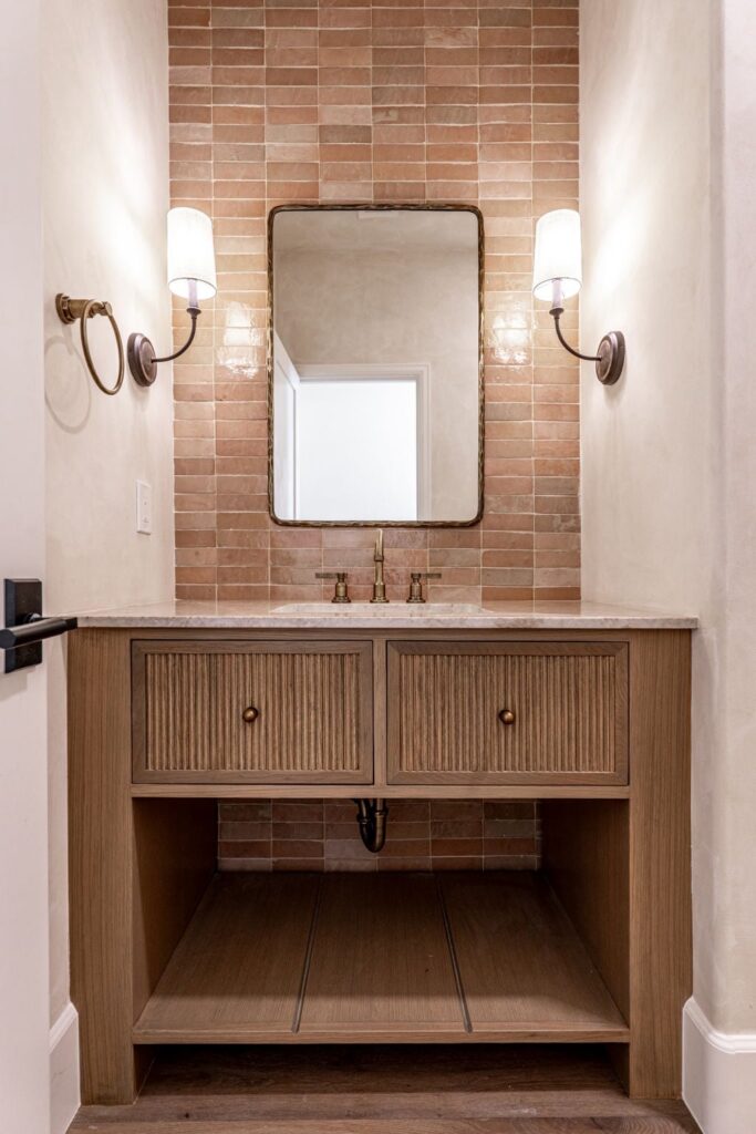 a bathroom vanity with a rectangular mirror, two wall sconces, a marble countertop, bronze fixtures, and a tiled backsplash in earth tones.
