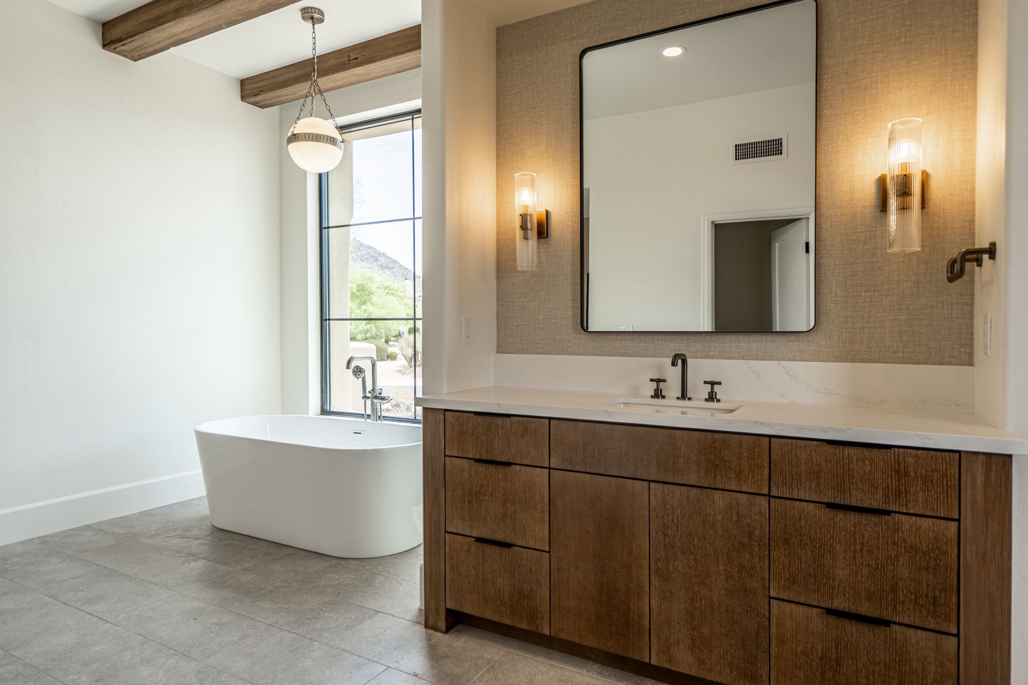 modern bathroom with a freestanding tub by a large window, wooden vanity with double sinks, wall mounted mirror, and two sconces.