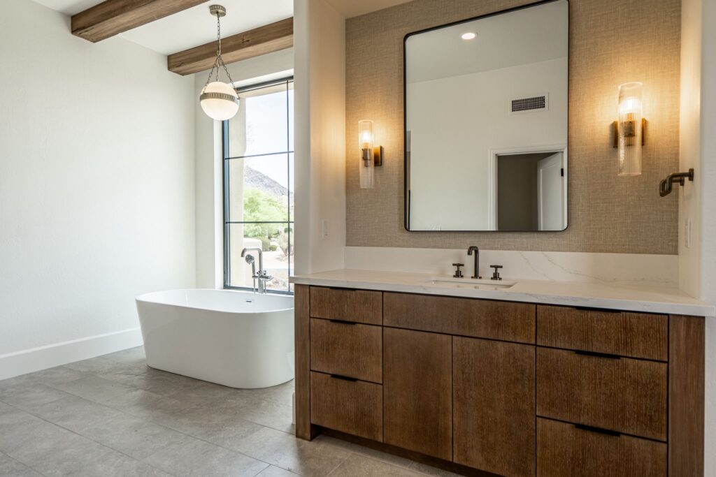 Modern bathroom with a freestanding tub by a large window, wooden vanity with double sinks, wall-mounted mirror, and two sconces.