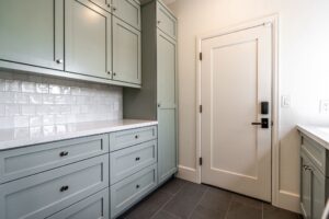 A kitchen corner with sage green cabinets, white tile backsplash, white countertop, gray tiled floor, and a closed white door with a black handle.
