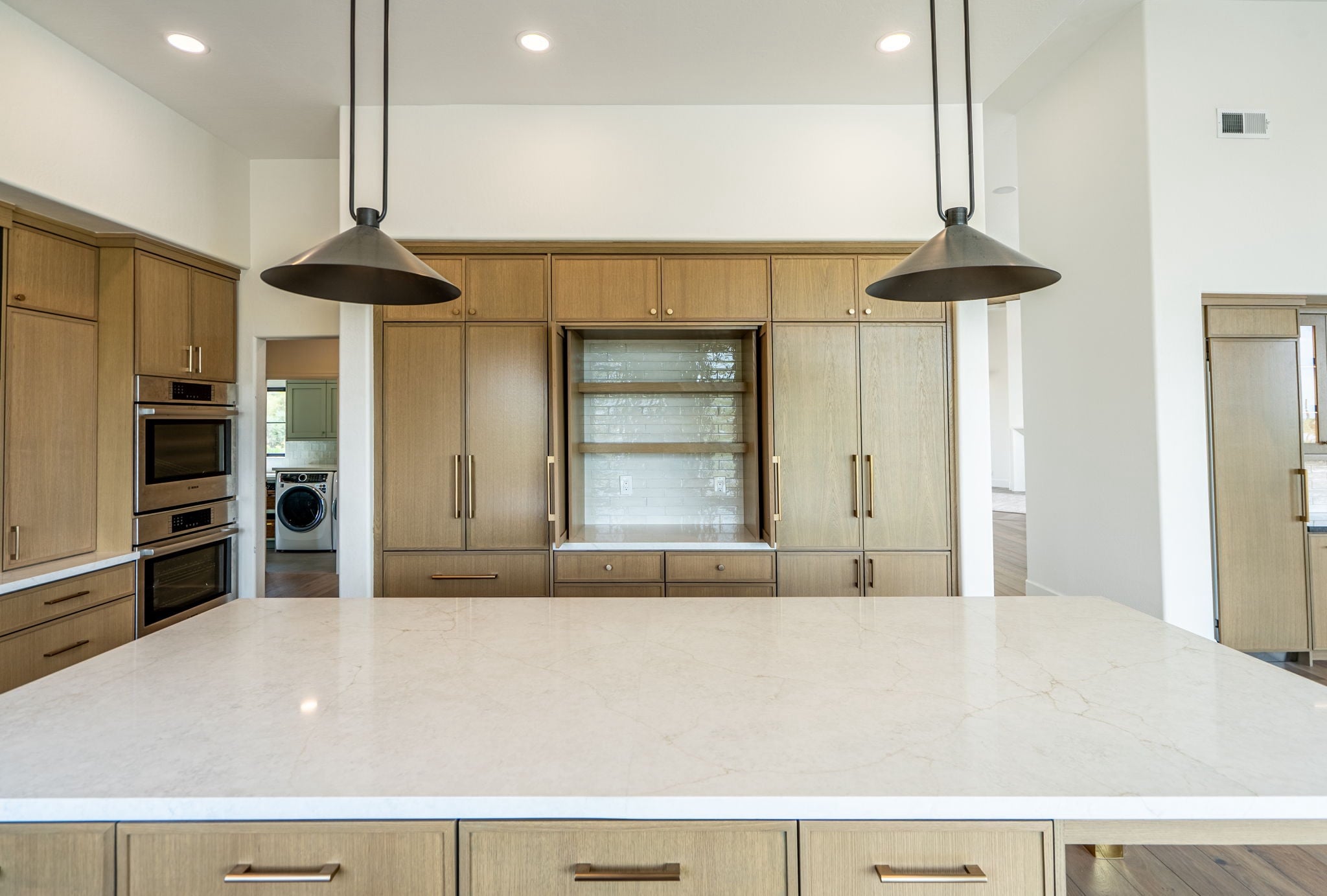 modern kitchen with a large marble island, wooden cabinets, built in appliances, and two black pendant lights. laundry room visible through an open doorway to the left.