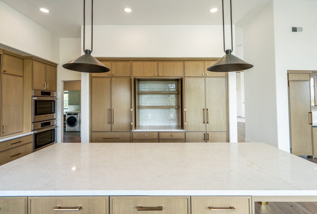 Modern kitchen with a large marble island, wooden cabinets, built-in appliances, and two black pendant lights. Laundry room visible through an open doorway to the left.