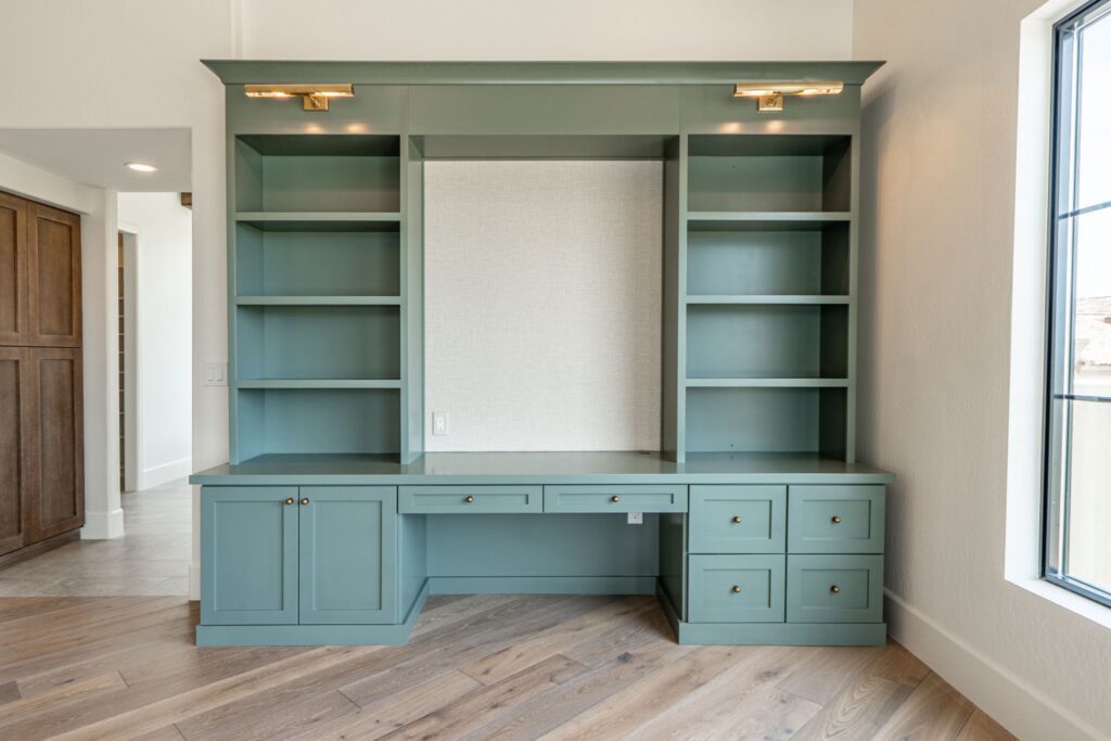 Built-in green desk with drawers and open shelves against a wall, featuring wood flooring and a nearby window letting in natural light.