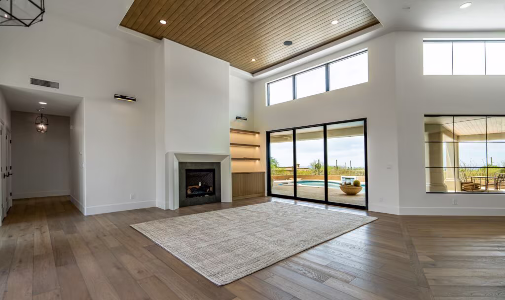 Spacious living room with wood flooring, large windows, fireplace, built-in shelves, and sliding glass doors leading to an outdoor patio and pool area.