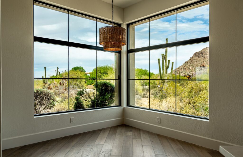 a corner room with large windows showing a desert landscape with cacti, bushes, and hills. a woven pendant light hangs from the ceiling above light wood flooring.