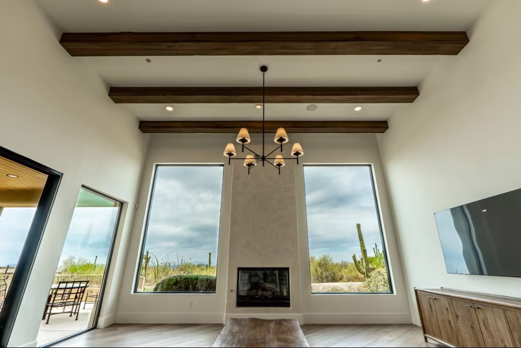 Modern living room with wood ceiling beams, a central fireplace, large windows showing a desert view, a chandelier, a flat-screen TV, and wooden furniture.
