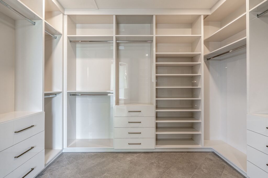 Empty walk-in closet with built-in shelves, drawers, and hanging rods, featuring a modern, glossy white finish and gray tiled floor.