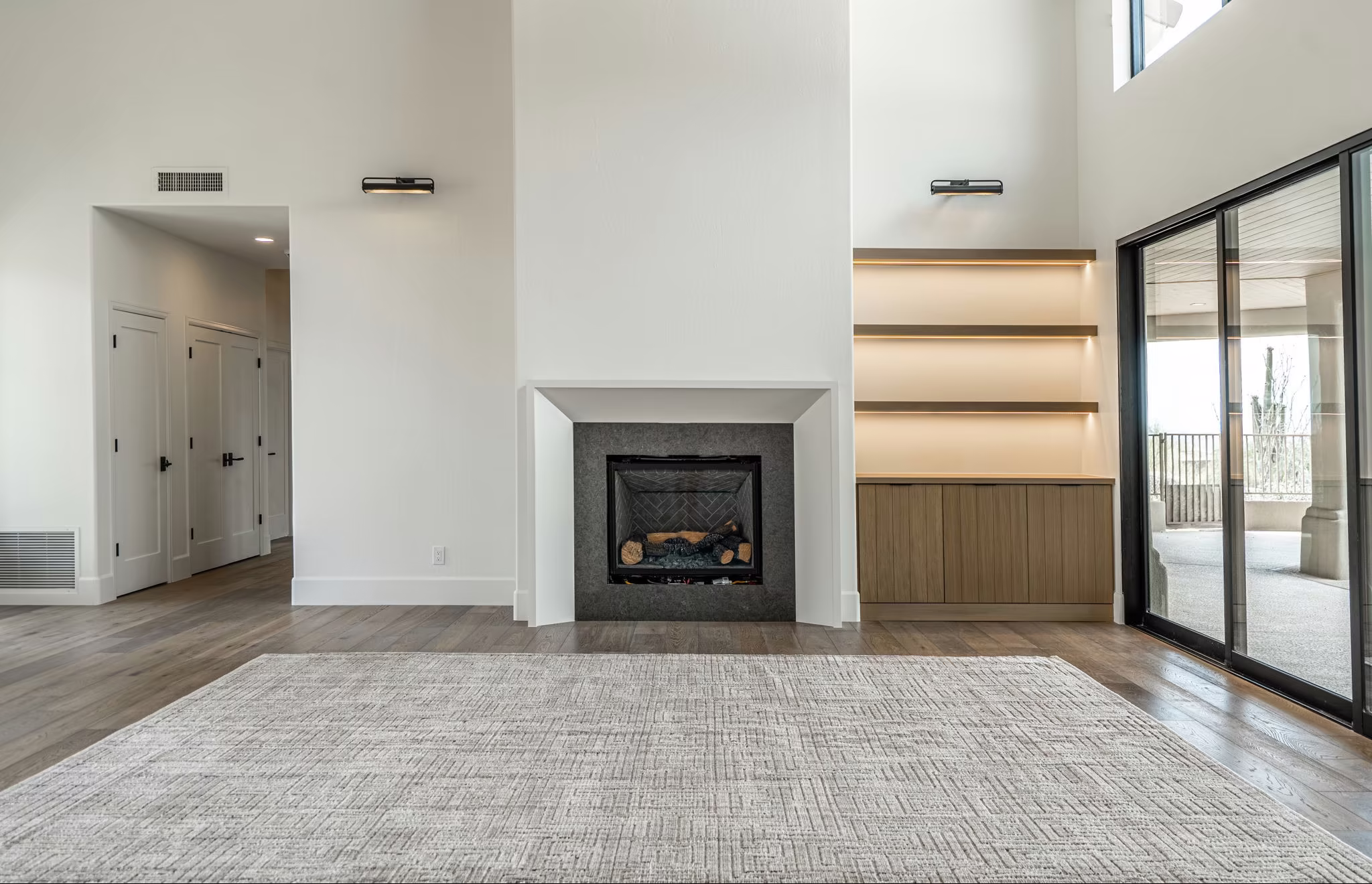 A modern living room with a fireplace, built-in shelves, large rug, wood flooring, double doors, and sliding glass doors to a balcony.