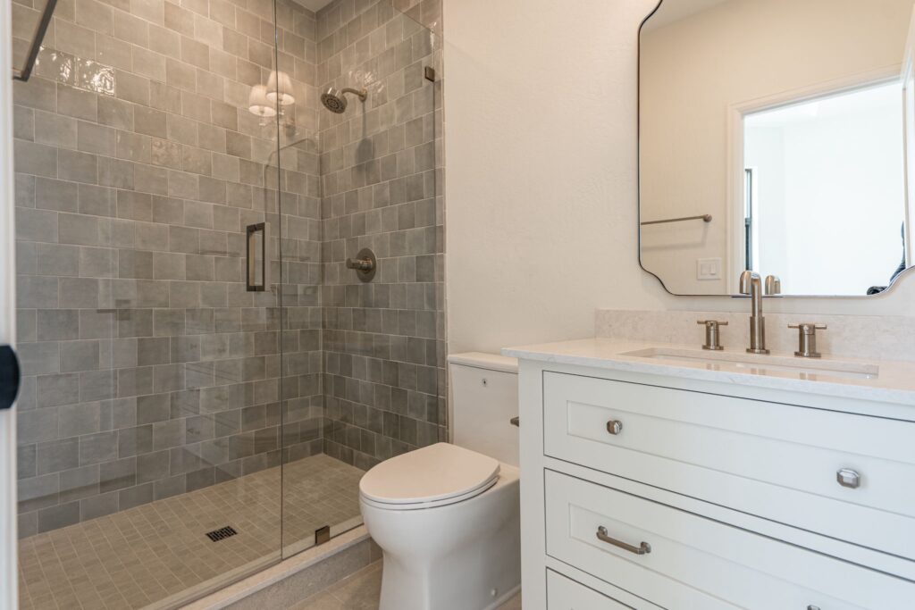 Modern bathroom with a glass-enclosed shower, gray tile walls, a white vanity with a sink and brass fixtures, and a toilet.