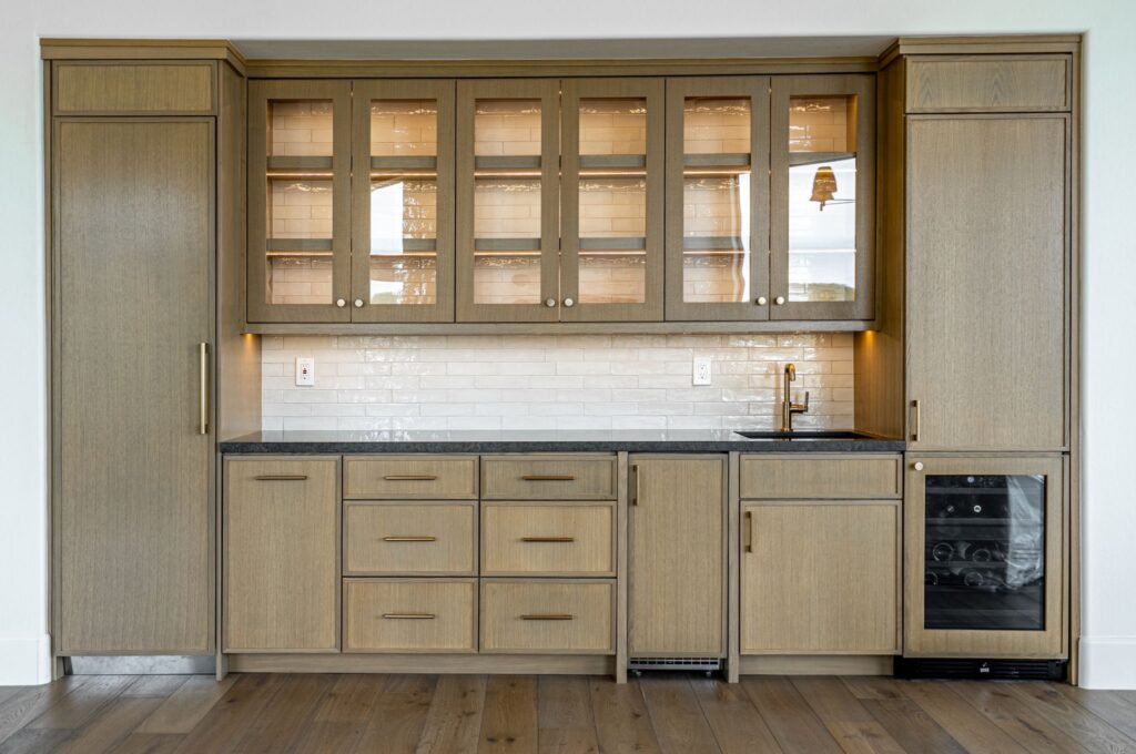 Modern kitchen with light wood cabinets, glass upper doors, black countertop, white subway tile backsplash, built-in wine cooler, and under-cabinet lighting.