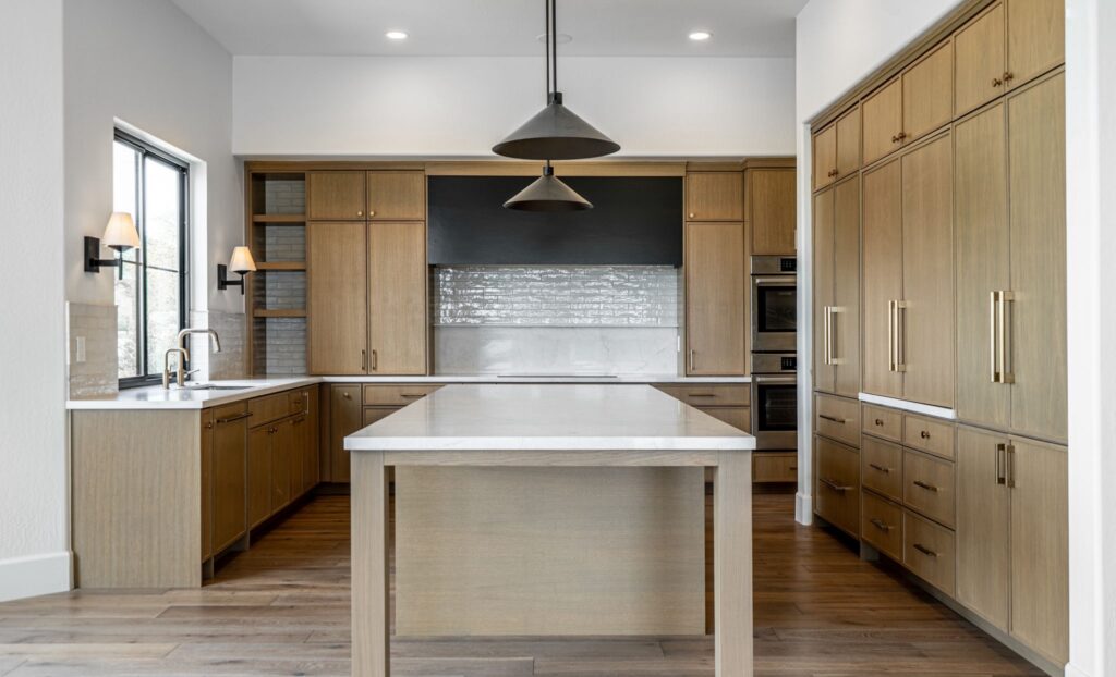 Modern kitchen with wood cabinets, white countertops, a large central island, pendant lights, built-in ovens, and a window above the sink.