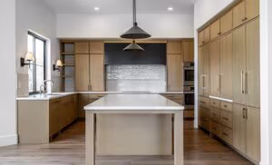 Modern kitchen with wood cabinets, white countertops, a large central island, pendant lights, built-in ovens, and a window above the sink.