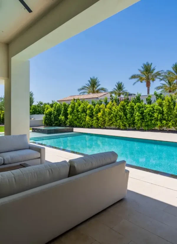 Covered patio overlooking a clear blue swimming pool, with beige tile flooring and two white sofas. The area is surrounded by lush green trees and palm trees under a clear blue sky. This serene setting is crafted by the best home remodeling contractors near me, showcasing modern architecture through glass doors on the left.