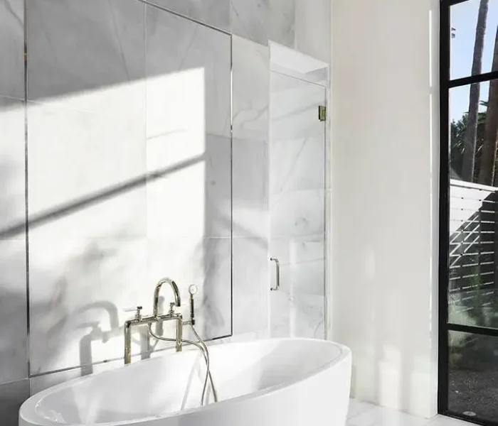 During a recent bathroom renovation, a freestanding white oval bathtub was expertly placed against a marble-tiled wall. A gold faucet and modern ceiling light add elegance. Sunlight streams through a large window with a black frame, creating shadows on the floor, enhancing the luxurious ambiance.