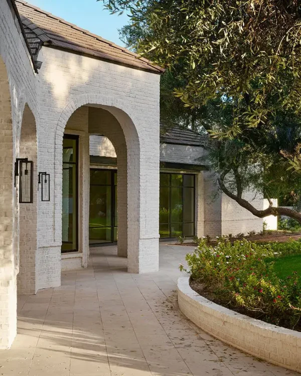 A modern house exterior with a white brick facade and large arched doorways complements the recently completed kitchen remodeling. Tall glass windows overlook a patio with beige tiles. Manicured bushes and a grassy lawn accentuate the scene, while trees provide ample shade.