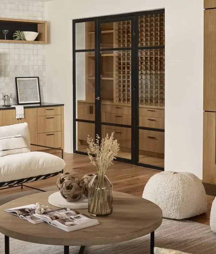 A modern living room features a cozy sitting area with two white cushioned chairs and a wood coffee table adorned with books and dried plants. Beige poufs sit on the floor, while a wooden cabinet and fireplace line the wall, showcasing the impeccable touch of a general contractor in Scottsdale.