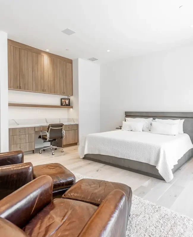 A minimalist bedroom crafted by the best home remodeling contractors near me features a large bed with white bedding, flanked by small round side tables. Two brown leather armchairs face the bed, and a built-in wooden desk is seamlessly integrated into the wall against light wood flooring and white walls.