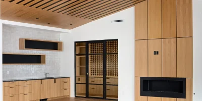 A modern room with light wood flooring and a ceiling of horizontal slats showcases impeccable kitchen remodeling. Built-in wooden cabinetry surrounds the fireplace, while a kitchenette with a sink, open shelves, and tiled backsplash enhances functionality. Glass doors lead to wine storage with honeycomb shelves.